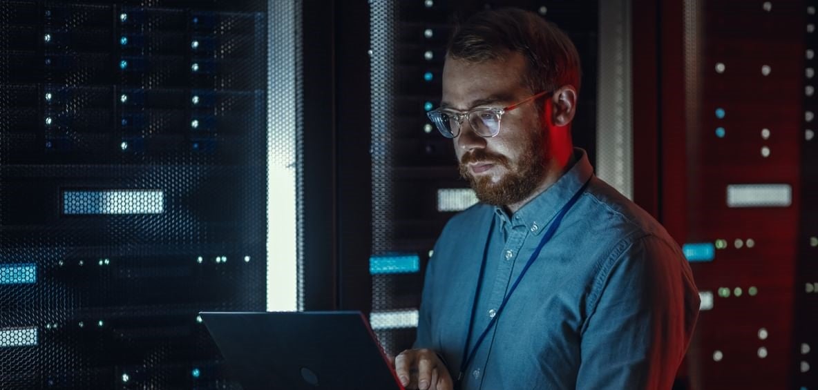 Bearded IT Specialist in Glasses is Working on Laptop in Data Center while Standing Near Server Rack