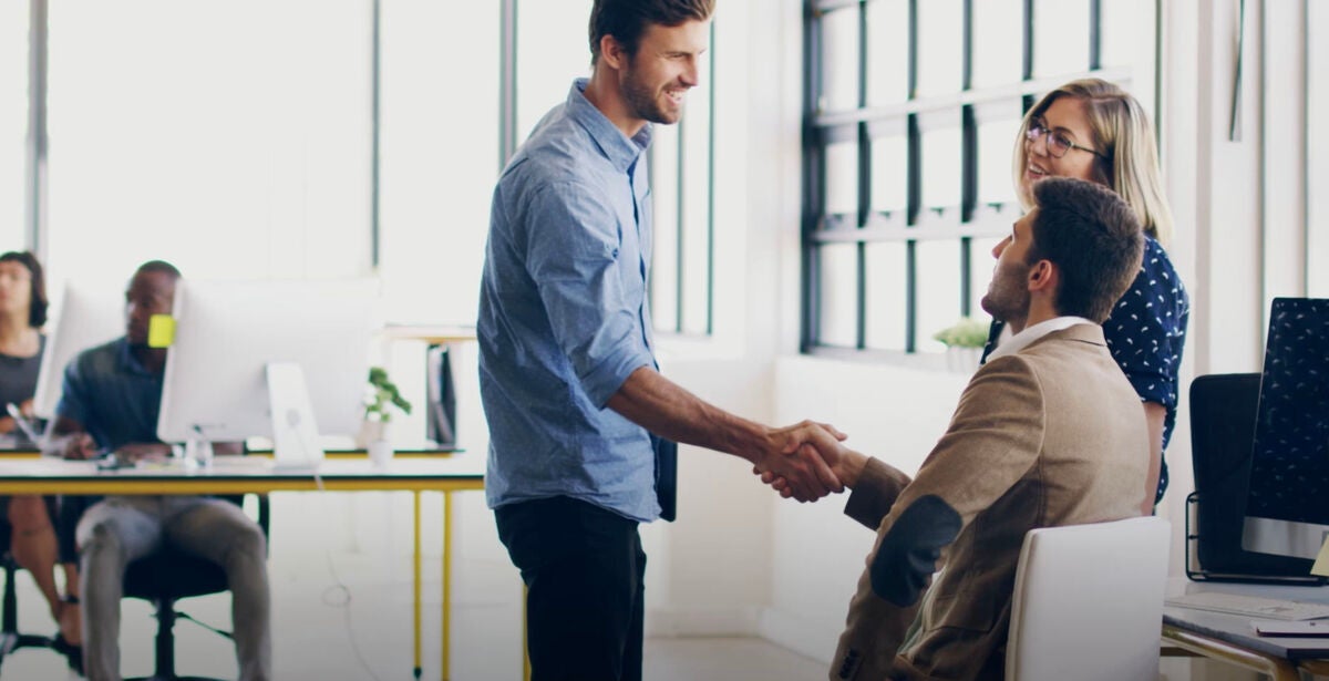 A man in a brown jacket sitting down at a computer shaking hands with another who's standing up, thinking a masters degree really helps with expanding his network.