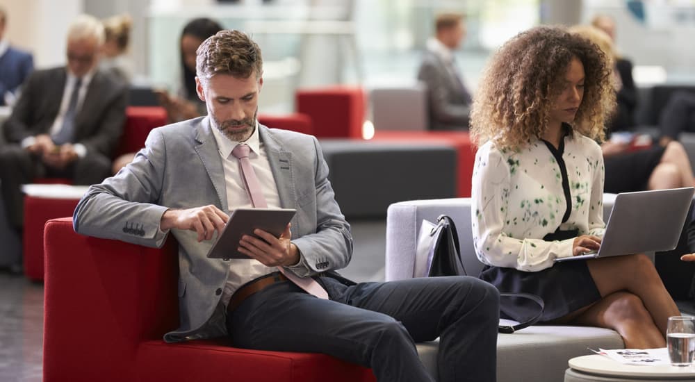 Male and female business professionals sat on a sofa reading on electronic devices.