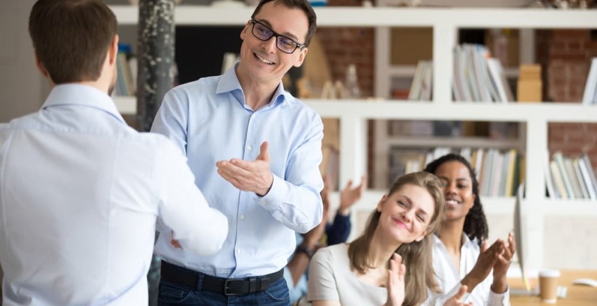 Two business men shaking hands at a business meeting, while other colleagues applaud them.